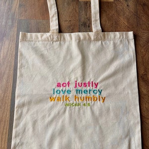 May include: A white canvas tote bag with the text "act justly love mercy walk humbly Micah 6:8" embroidered in colorful thread.