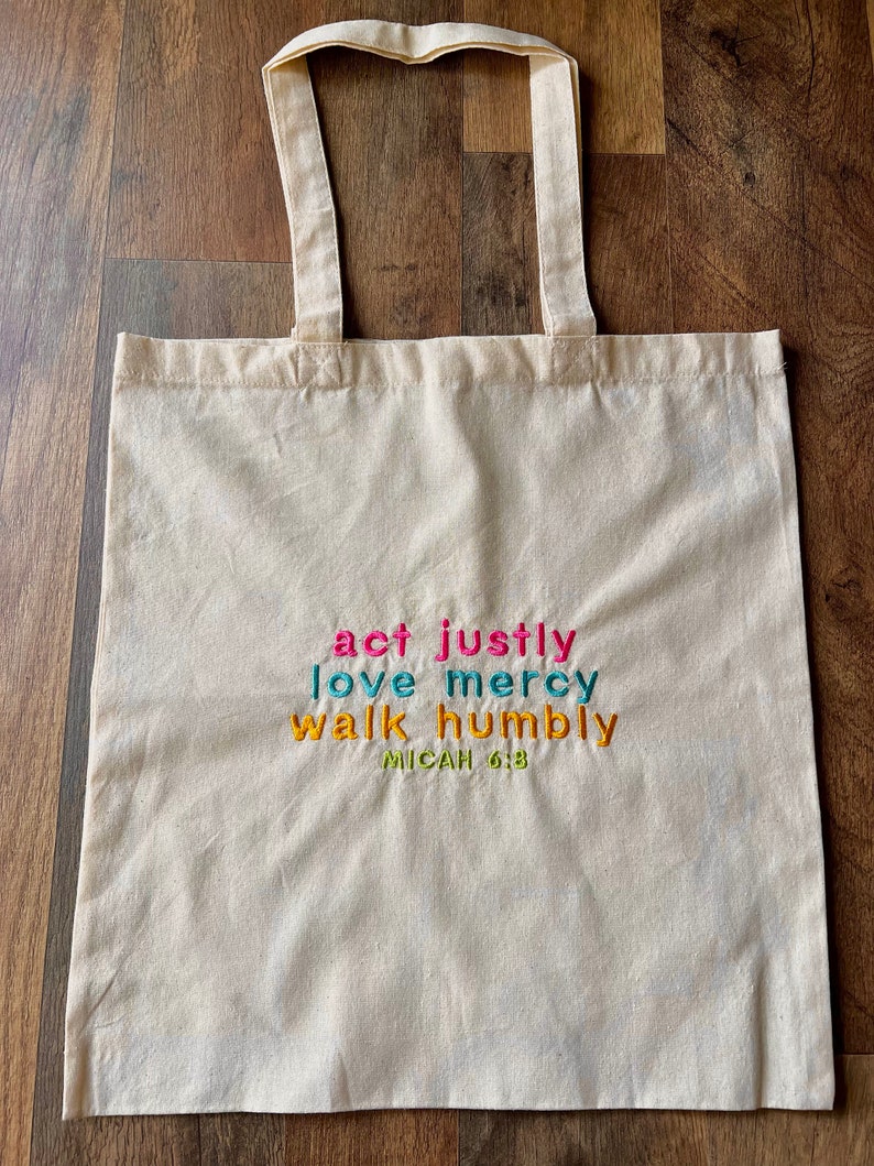 May include: A white canvas tote bag with the text "act justly love mercy walk humbly Micah 6:8" embroidered in colorful thread.