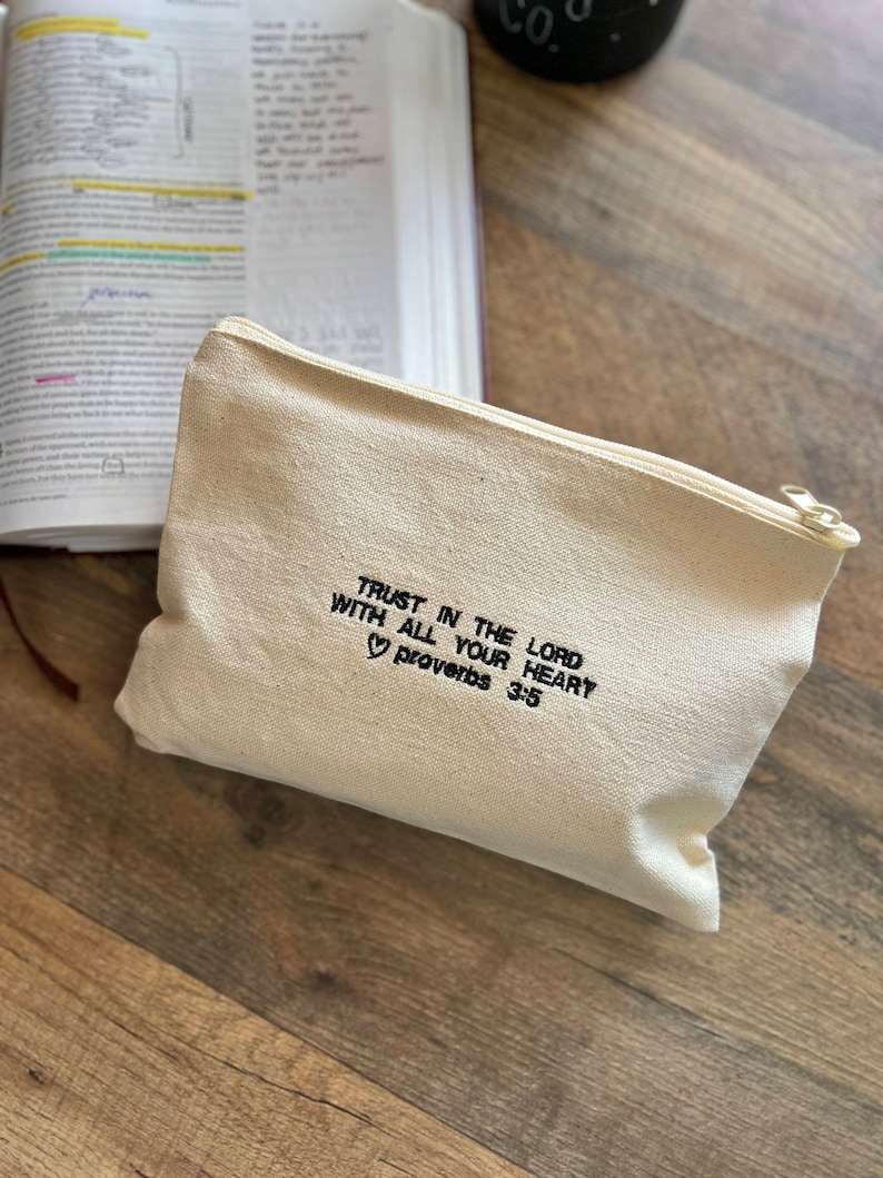 Peut inclure: Une pochette zipp&eacute;e en toile blanche avec le texte "TRUST IN THE LORD WITH ALL YOUR HEART ♡ Proverbs 3:5" brod&eacute; en noir.