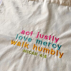 May include: A white canvas tote bag with the words "act justly, love mercy, walk humbly" embroidered in colorful thread. The text "Micah 6:8" is also embroidered in green thread below the main text.
