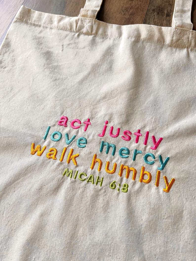 May include: A white canvas tote bag with the words "act justly, love mercy, walk humbly" embroidered in colorful thread. The text "Micah 6:8" is also embroidered in green thread below the main text.