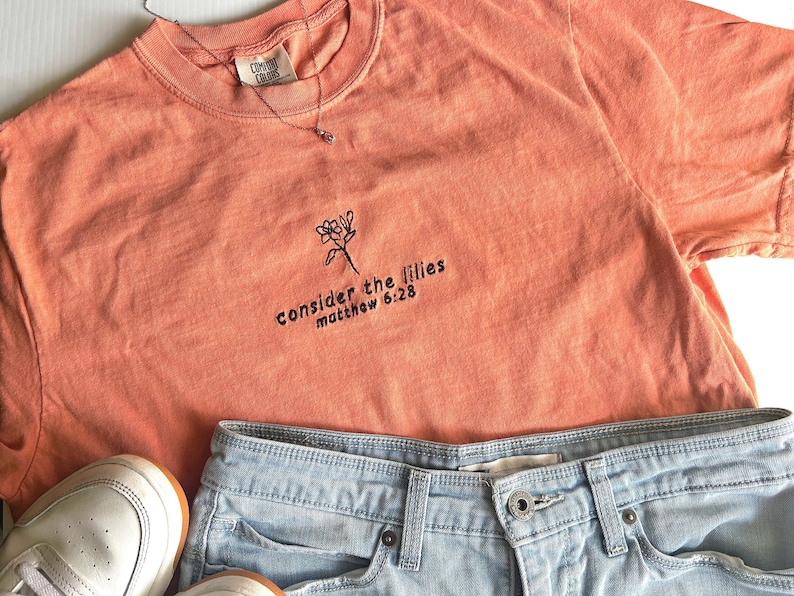 May include: A coral orange t-shirt with a black embroidered flower and the text "consider the lilies matthew 6:28".