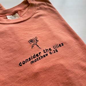 May include: A coral colored t-shirt with black embroidery that reads "consider the lilies matthew 6:28".