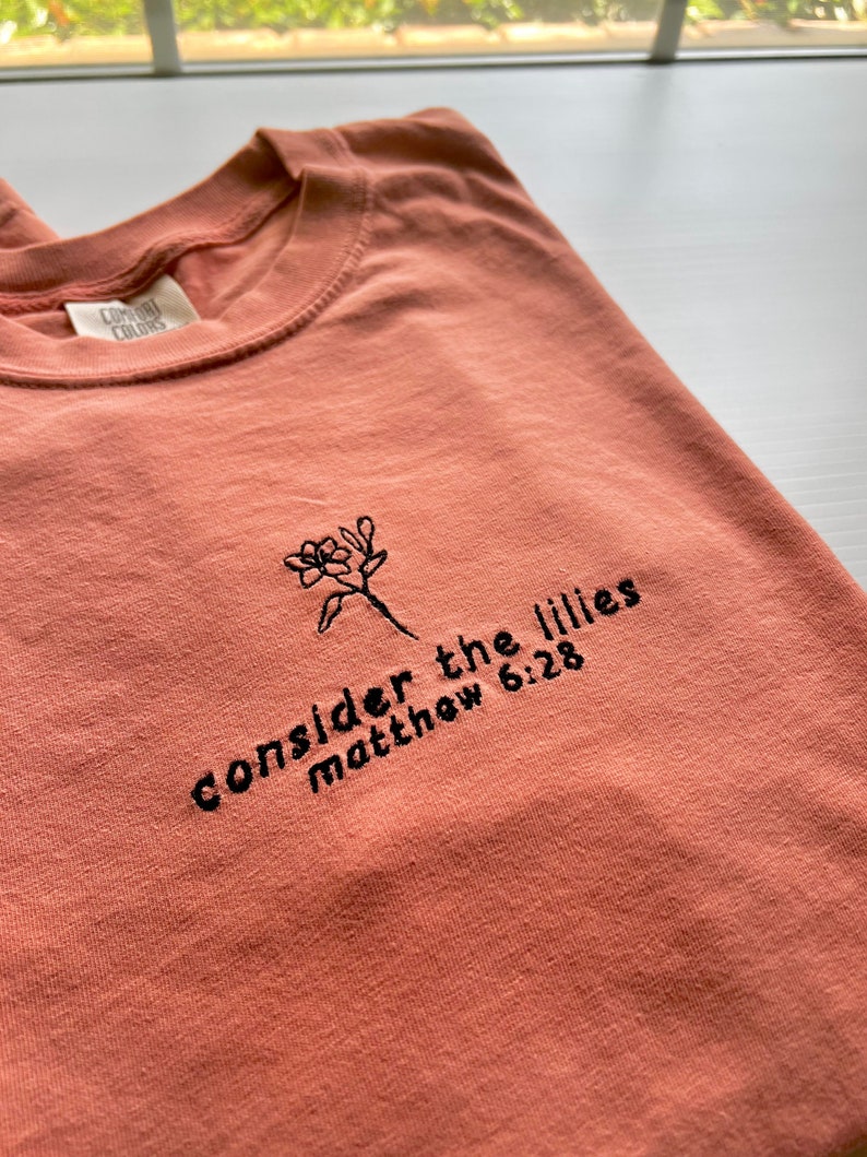 May include: A coral colored t-shirt with black embroidery that reads "consider the lilies matthew 6:28".