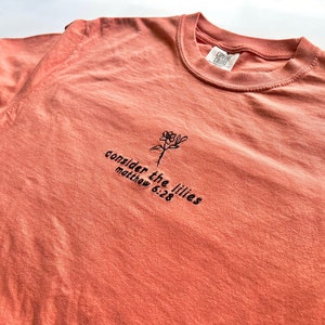 May include: A coral colored t-shirt with a black embroidered flower and the text "consider the lilies matthew 6:28"