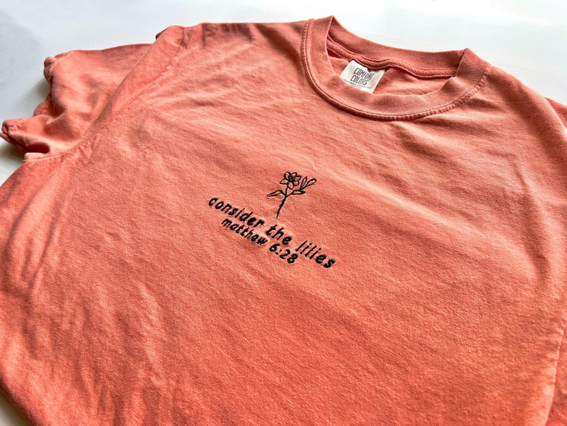 May include: A coral colored t-shirt with a black embroidered flower and the text "consider the lilies matthew 6:28"