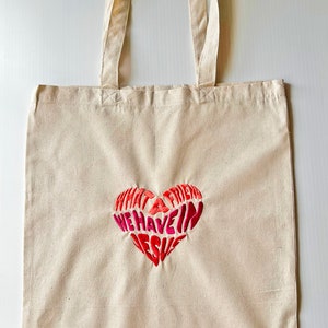 Op de afbeelding: Een beige canvas draagtas met een rood en roze geborduurd hart waarop staat "Wat een vriend hebben we in Jezus".
