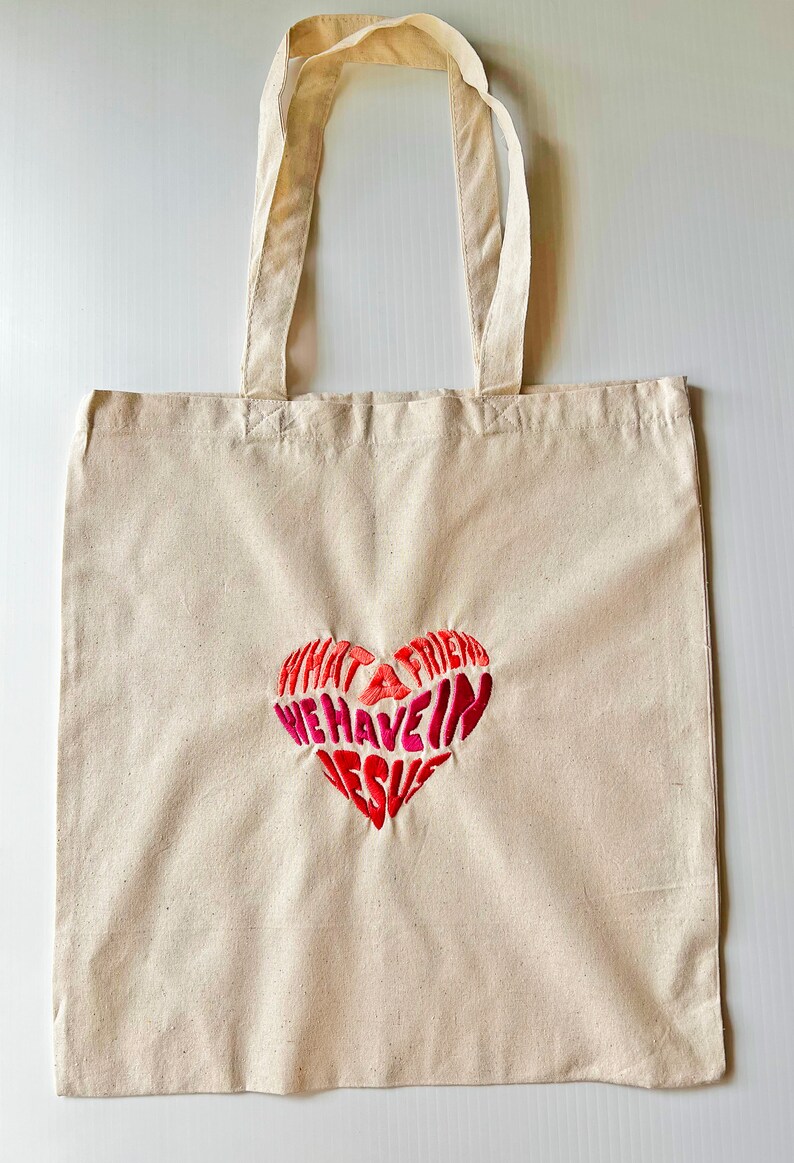 Op de afbeelding: Een beige canvas draagtas met een rood en roze geborduurd hart waarop staat "Wat een vriend hebben we in Jezus".