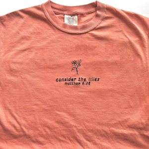 May include: A coral colored t-shirt with black embroidery of a flower and the text "consider the lilies matthew 6:28"