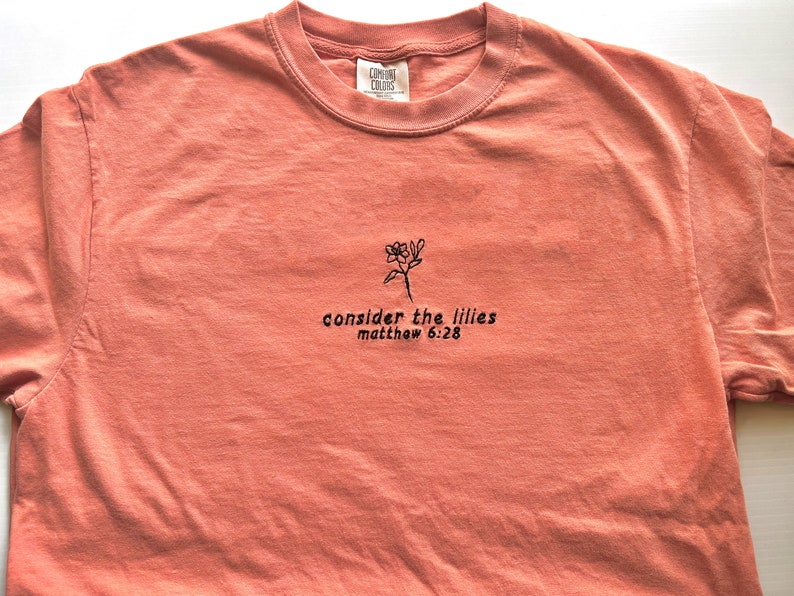 May include: A coral colored t-shirt with black embroidery of a flower and the text "consider the lilies matthew 6:28"