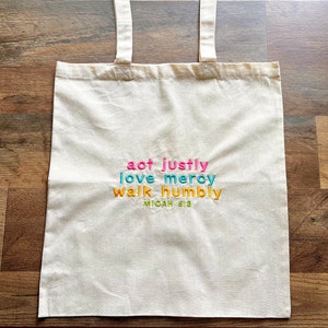 May include: A white canvas tote bag with the text "act justly love mercy walk humbly Micah 6:8" embroidered in colorful thread.
