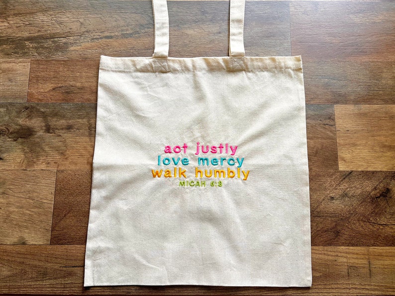 May include: A white canvas tote bag with the text "act justly love mercy walk humbly Micah 6:8" embroidered in colorful thread.