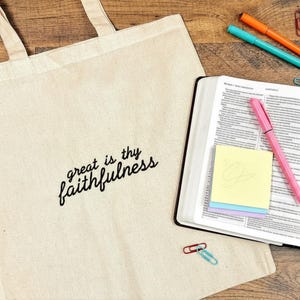 Puede incluir: Una bolsa de lona beige con el texto "great is thy faithfulness" en escritura negra. Un libro abierto con un bol&iacute;grafo rosa, notas adhesivas y marcadores de colores est&aacute;n al lado de la bolsa. Clips de papel est&aacute;n esparcidos sobre la superficie de madera.