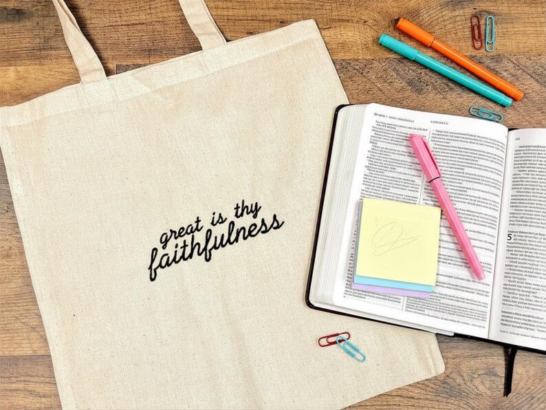 Puede incluir: Una bolsa de lona beige con el texto "great is thy faithfulness" en escritura negra. Un libro abierto con un bol&iacute;grafo rosa, notas adhesivas y marcadores de colores est&aacute;n al lado de la bolsa. Clips de papel est&aacute;n esparcidos sobre la superficie de madera.