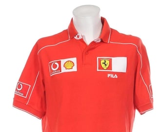 Può includere: Polo rosso a maniche corte con bordini e loghi bianchi. I loghi includono Vodafone, Shell, Ferrari e Fila.