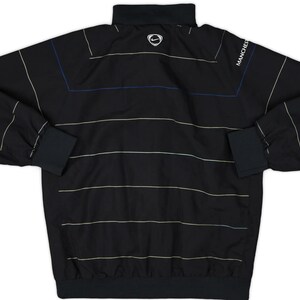 K&ouml;nnte beinhalten: Schwarze Langarm-Sportjacke mit horizontalen wei&szlig;en und beigen Streifen. Mit schwarzem Kragen, Manschetten und Saum. Das Nike-Logo ist auf dem oberen R&uuml;cken sichtbar, und das Wort "MANCHESTER" ist auf dem &Auml;rmel aufgedruckt.