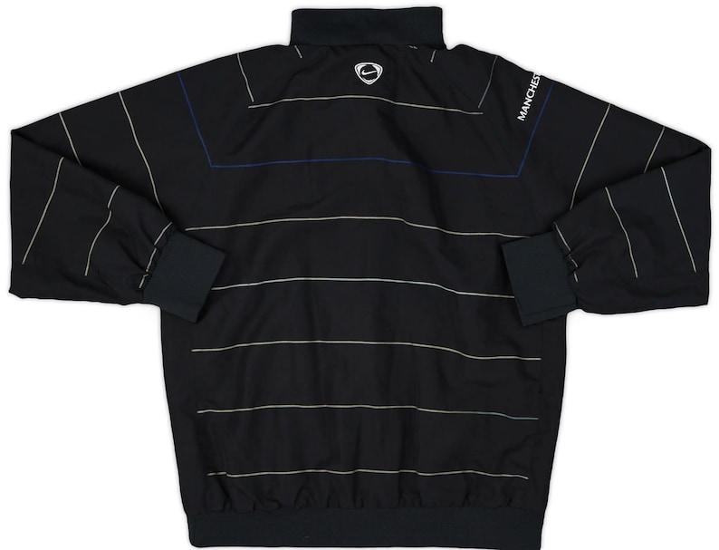 K&ouml;nnte beinhalten: Schwarze Langarm-Sportjacke mit horizontalen wei&szlig;en und beigen Streifen. Mit schwarzem Kragen, Manschetten und Saum. Das Nike-Logo ist auf dem oberen R&uuml;cken sichtbar, und das Wort "MANCHESTER" ist auf dem &Auml;rmel aufgedruckt.