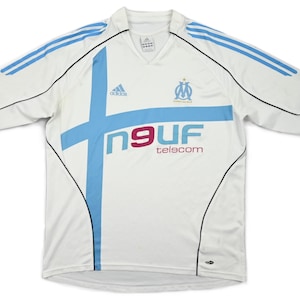 May include: White soccer jersey with light blue accents and the Adidas logo. The jersey features a light blue cross design and the text "n9UF telscom" in red and blue. The sleeves have light blue stripes and black trim.
