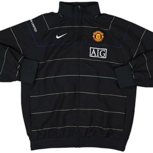 K&ouml;nnte beinhalten: Schwarze Jacke mit Rei&szlig;verschluss und wei&szlig;en horizontalen Streifen. Mit Nike-Logo, Vereinswappen und AIG-Logo. Auf dem linken &Auml;rmel steht "MANCHESTER UNITED". Die Jacke hat einen Stehkragen und schwarze B&uuml;ndchen.