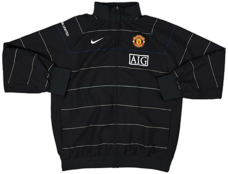 K&ouml;nnte beinhalten: Schwarze Jacke mit Rei&szlig;verschluss und wei&szlig;en horizontalen Streifen. Mit Nike-Logo, Vereinswappen und AIG-Logo. Auf dem linken &Auml;rmel steht "MANCHESTER UNITED". Die Jacke hat einen Stehkragen und schwarze B&uuml;ndchen.
