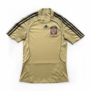 May include: A gold short-sleeved athletic jersey featuring three black stripes along each shoulder and sleeve. The shirt displays a black Adidas logo on the center chest and the Spanish national football team crest on the upper left chest area.