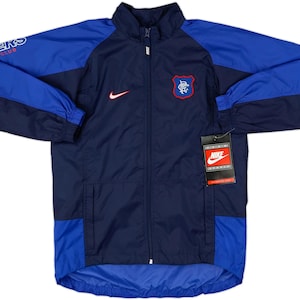 May include: A navy and royal blue Nike jacket with a full zip. The jacket displays the Rangers Football Club emblem on the chest and the Nike swoosh. The sleeves have the text "RANGERS FOOTBALL CLUB".