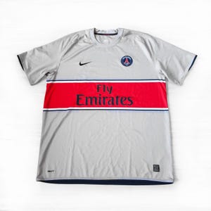 May include: Grey short-sleeved football shirt with a red stripe across the chest, displaying the text "Fly Emirates". Includes a team badge and a Nike logo. The shirt has navy blue trim.