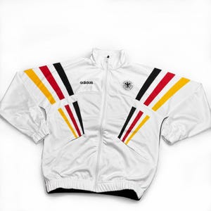 May include: White Adidas track jacket with black, red, and yellow stripes on the sleeves. Features a full-length zipper, the Adidas logo, and a German national team emblem. The jacket has a high collar and a sporty design.