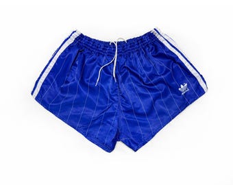 May include: Royal blue athletic shorts with white vertical stripes and white side stripes. The shorts have a drawstring waist and the Adidas logo on the leg. A vintage style.