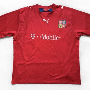 Peut inclure: Un maillot de sport rouge à manches courtes avec un logo Puma blanc sur la poitrine et les épaules. L'avant affiche le texte blanc T-Mobile et un écusson coloré représentant un lion sur le côté droit. Le col possède une doublure intérieure bleue.