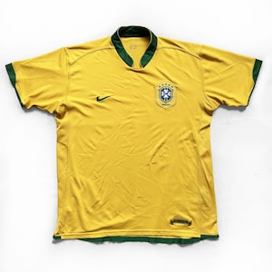 Puede incluir: Una camiseta de fútbol amarilla con ribetes verdes en el cuello y las mangas. La camiseta presenta un logotipo de Nike y el emblema de la Confederación Brasileña de Fútbol. La camiseta es de material ligero.