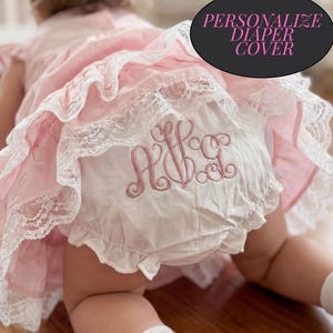 May include: A white diaper cover featuring pink embroidered monogram lettering and ruffled lace trim, layered under a pink dress with matching lace details. The text PERSONALIZE DIAPER COVER is visible in a black oval graphic overlay.