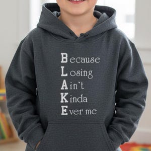 Può includere: Una felpa con cappuccio grigio scuro con tasca frontale e cappuccio. Il testo "Because Losing Ain't Kinda Ever me" è stampato in bianco. La felpa con cappuccio è indossata da un bambino.