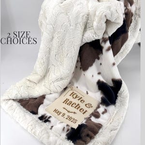 May include: A soft, cream-coloured blanket with a cowhide pattern in brown and black. The blanket has a textured surface and a cream-coloured border. A rectangular patch is embroidered with the names "Kyle & Rachel" and the date "May 9, 2025". The text "2 SIZE CHOICES" is also visible.