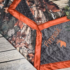 May include: A gray and black baby blanket with a camouflage print on one side and a gray side with an orange satin border. The gray side has the name "Finnick" embroidered on it with a deer head design.