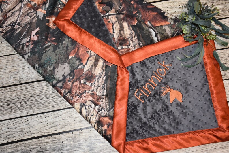 May include: A gray and black baby blanket with a camouflage print on one side and a gray side with an orange satin border. The gray side has the name "Finnick" embroidered on it with a deer head design.