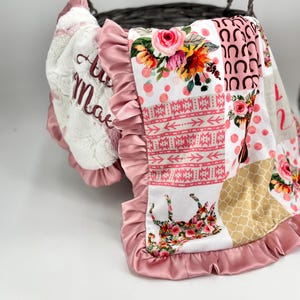 Op de afbeelding: Een babydeken met een bloemen- en hoefijzerpatroon. De deken heeft een roze ruchesrand en een wit gedeelte met de geborduurde naam "Ava Mae". De deken heeft een patchwork-ontwerp met verschillende patronen en kleuren.