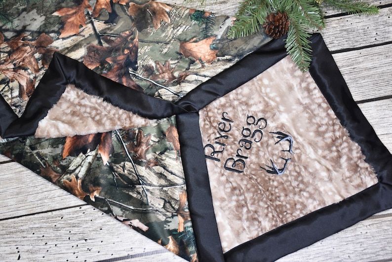 May include: A brown and green camouflage baby blanket with a soft, plush brown side. The blanket is personalized with the name "River Bragg" and a set of antlers.