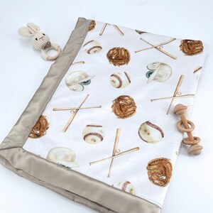 Op de afbeelding: Een witte babydeken met een honkbalthema, met honkballen, knuppels, handschoenen en helmen. De deken heeft een satijnen rand en wordt vergezeld door twee houten speeltjes: een konijnvormige bijtring en een ringratel.