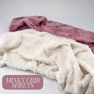 May include: Two minky crib sheets, one white and one pink, are shown folded and piled on top of each other. The sheets are made of a soft, plush fabric. The text "Minky Crib Sheets" is printed on a pink background.