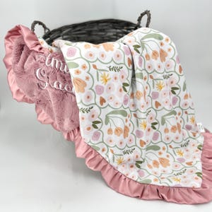 May include: A soft, plush blanket featuring a white background with a repeating pattern of pink, peach, and green flowers. One side is solid dusty rose faux fur with white embroidered text, finished with a matching dusty rose satin ruffle trim.