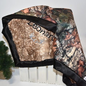 May include: A brown and tan faux fur baby blanket with a black satin border. The blanket has a camouflage print on one side and the name "Leonard" embroidered on the other side.