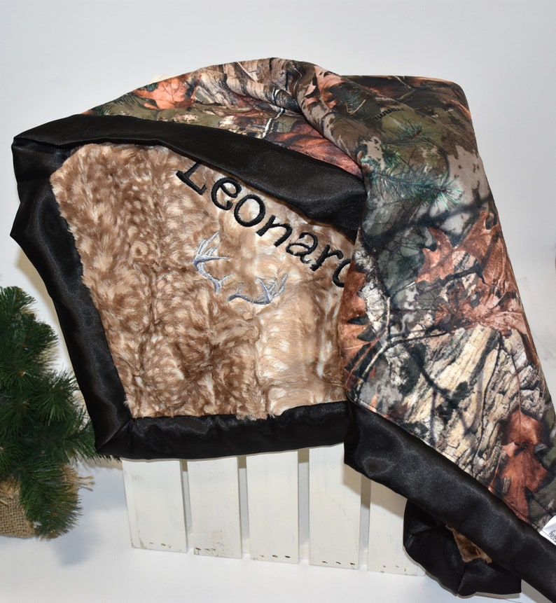 May include: A brown and tan faux fur baby blanket with a black satin border. The blanket has a camouflage print on one side and the name "Leonard" embroidered on the other side.
