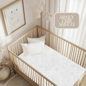 May include: A wooden crib with a white minky sheet and two white pillows. The sheet has a marble pattern. The text "MINKY CRIB SHEETS" is in a beige box in the upper right corner. A framed picture hangs on the wall.