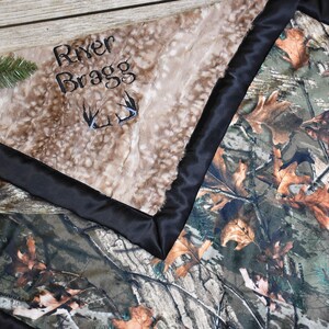 May include: A brown and tan faux fur baby blanket with a black satin border. The blanket has the name "River Bragg" embroidered on it with a pair of antlers. The other side of the blanket is a camouflage pattern.