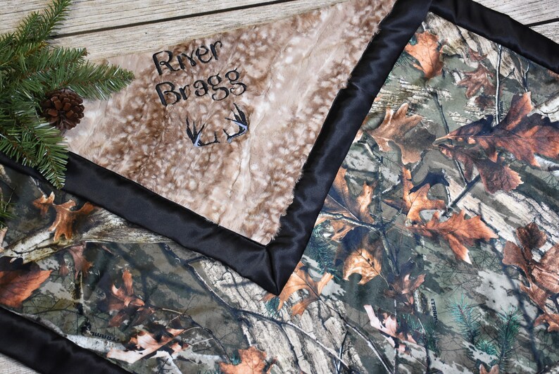 May include: A brown and tan faux fur baby blanket with a black satin border. The blanket has the name "River Bragg" embroidered on it with a pair of antlers. The other side of the blanket is a camouflage pattern.