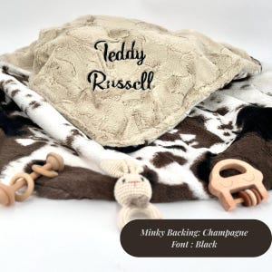 May include: A soft, beige minky baby blanket with the name "Teddy Russell" embroidered in black. The blanket has a cowhide pattern on the underside. A crochet bunny rattle and two wooden toys are also visible.