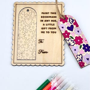 Puede incluir: Juego de marcapáginas y etiqueta de regalo de madera. El marcapáginas está pintado con flores rosas, moradas y blancas. La etiqueta dice "Pinta este marcapáginas en cualquier tono, un pequeño regalo de mí para ti". Incluye rotuladores.