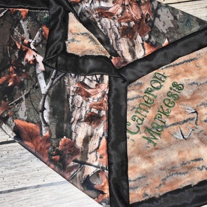 May include: A brown and tan camouflage fabric baby blanket with a black satin border. The blanket has a personalized embroidered name, "Cameron Markesis", in green thread with a green deer antler design.