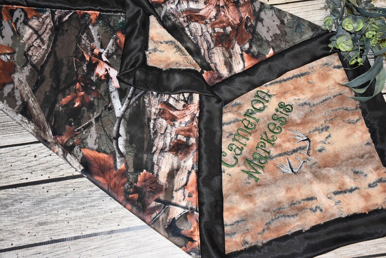 May include: A brown and tan camouflage fabric baby blanket with a black satin border. The blanket has a personalized embroidered name, "Cameron Markesis", in green thread with a green deer antler design.